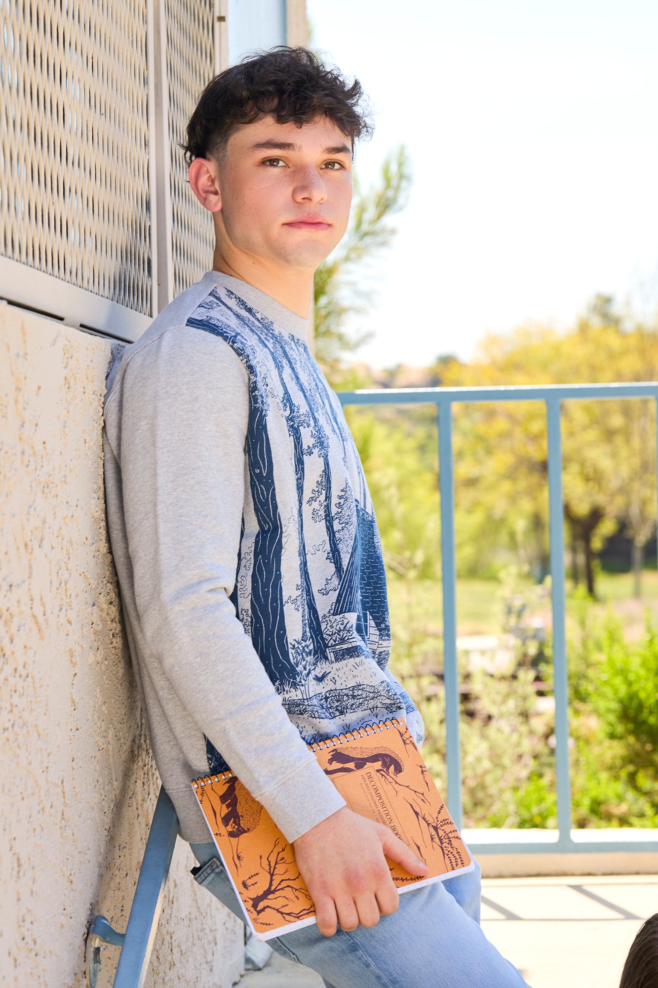 man wearing sweatshirt leaning against a wall holding notebook