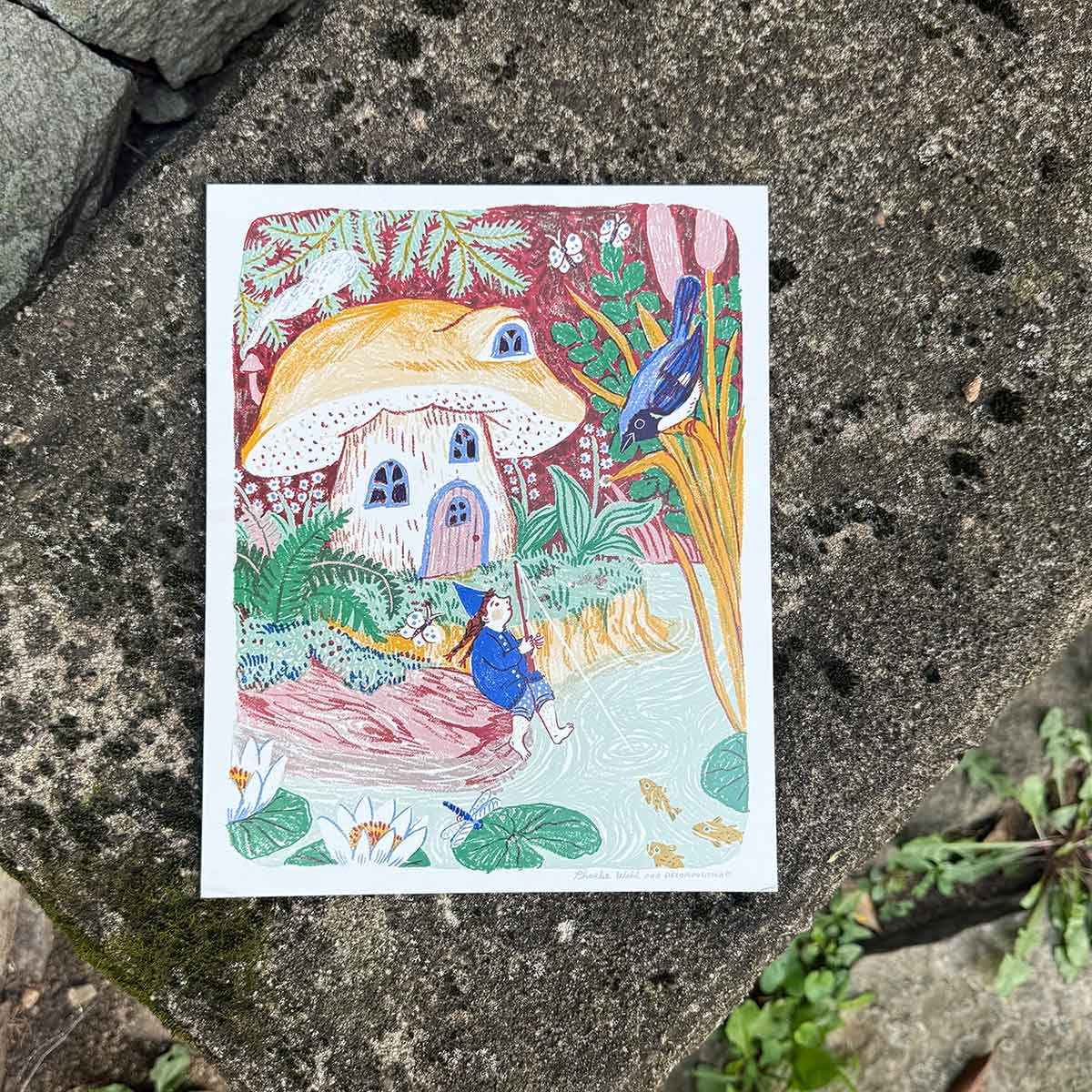 A rectangle art print featuring a whimsical forest illustration with a mushroom house, a figure in blue, and a bird, photographed flat on a textured gray stone surface.