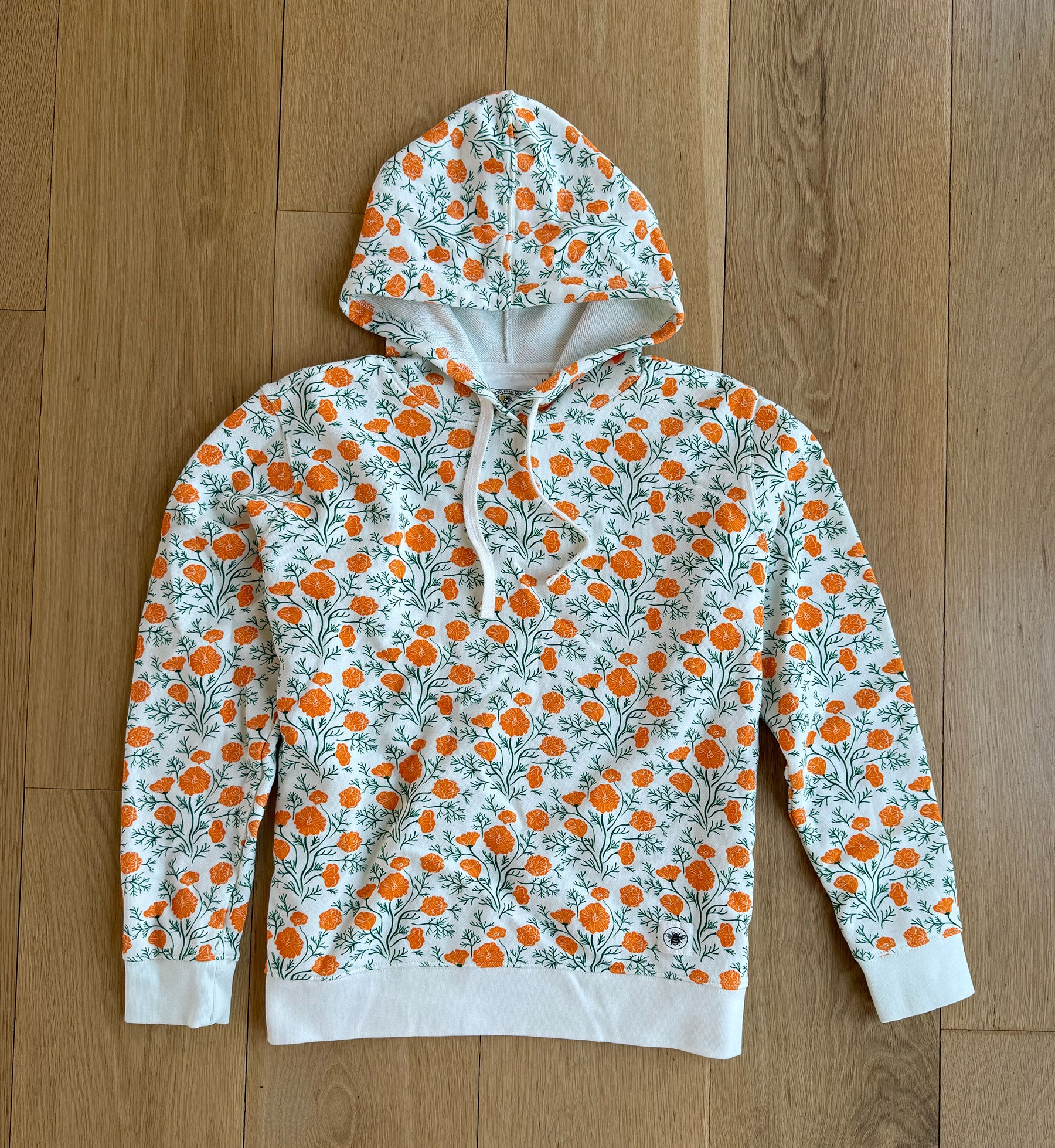 A hooded sweatshirt with a multicolored floral pattern, featuring orange flowers and green leaves on a white background, displayed on a wooden surface.