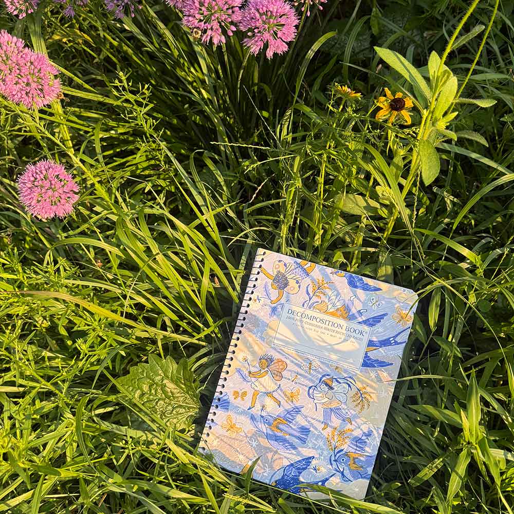 A spiral-bound notebook with a blue and white floral cover lies on green grass among pink flowers in an outdoor setting.
