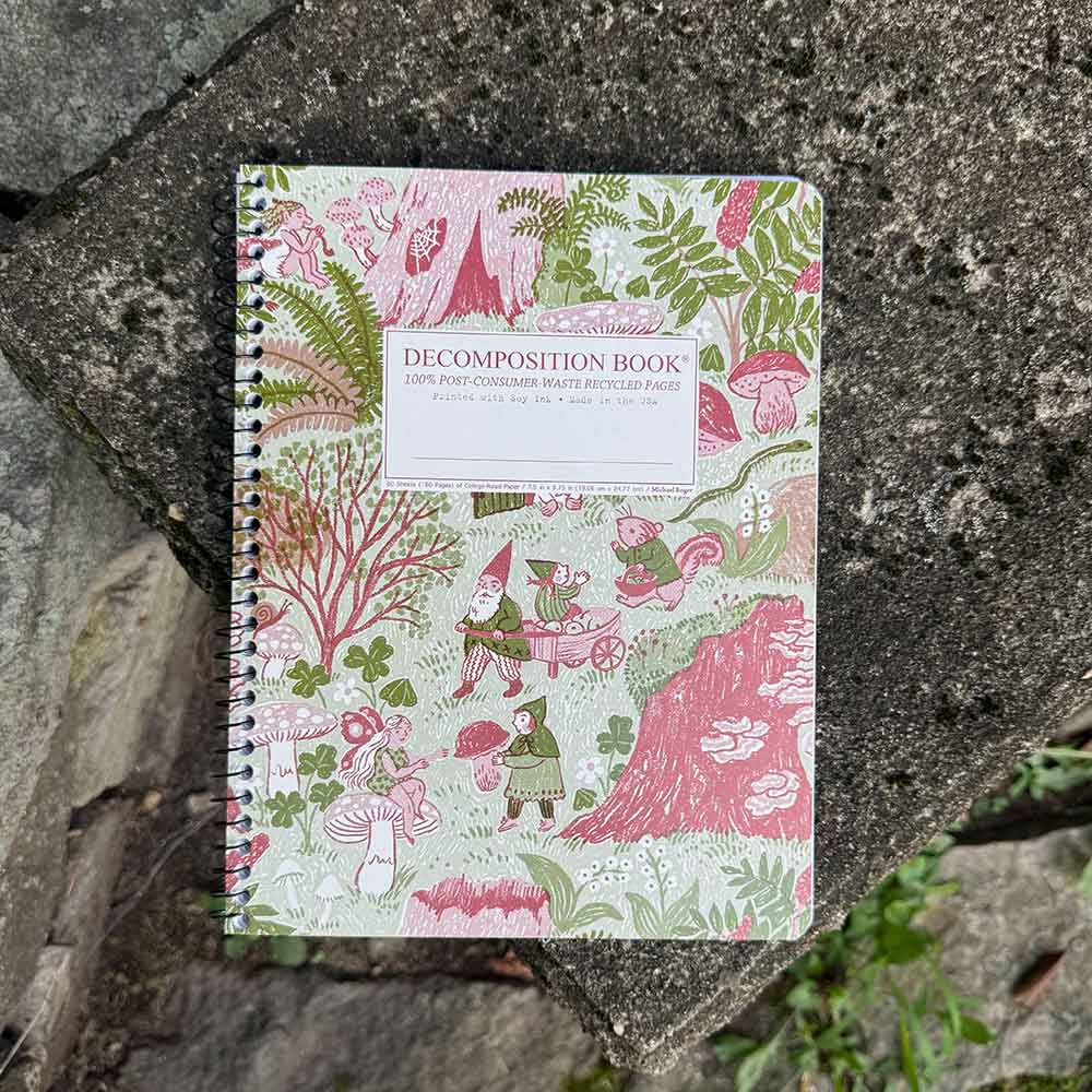 A spiral-bound decomposition book with a whimsical forest cover design in pink, green, and cream, photographed lying on a rough gray stone surface outdoors.