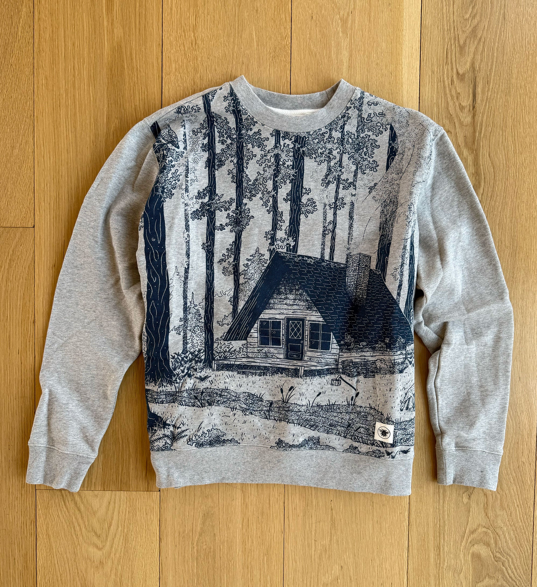 A gray crewneck sweatshirt with a printed design featuring a house in a forest setting.