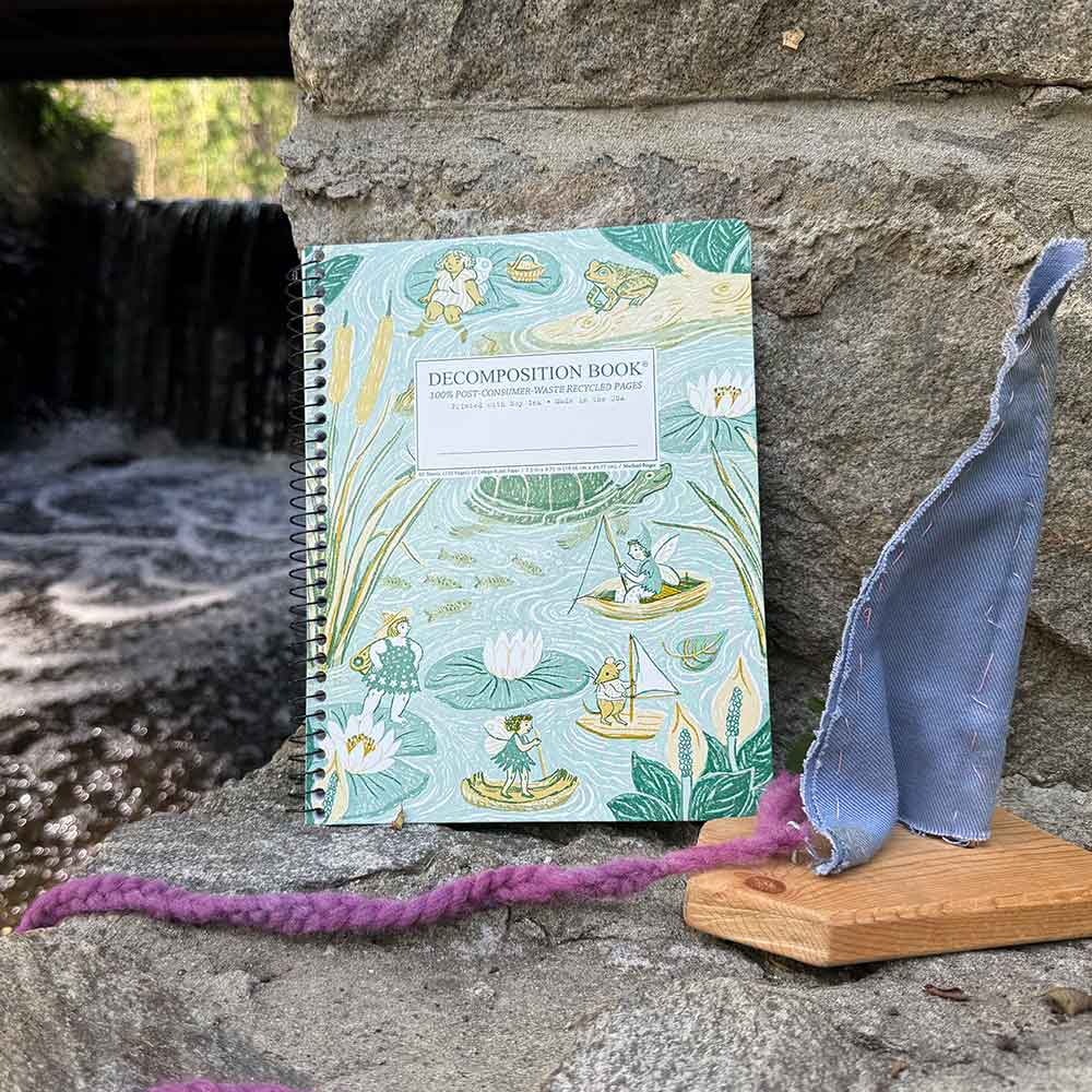 A spiral-bound composition book with a whimsical lily pond illustration stands on a stone ledge outdoors. The cover is light aqua with green and yellow botanical artwork and a white title label reading 'DECOMPOSITION BOOK'.